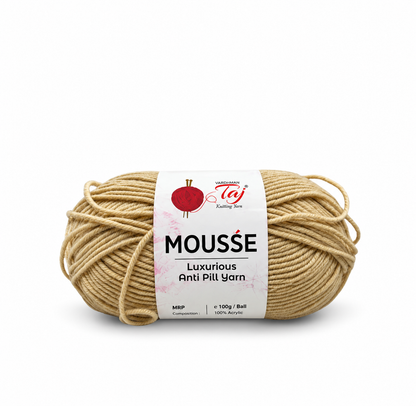 A skein of Mousse Luxurious Anti Pill Yarn in a light beige/tan color, wrapped with a white paper label featuring a red pom-pom logo and text, set against a plain white background.